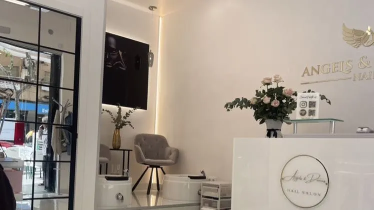 Interior del salón: recepción blanca con el logotipo redondo dorado, un jarrón de rosas y un cartel con códigos QR; al fondo, la zona de pedicura con dos sillones spa blancos, un sillón gris, un televisor y el logotipo con alas en la pared